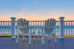 Two white beach chairs sitting on a deck in front of the beach.